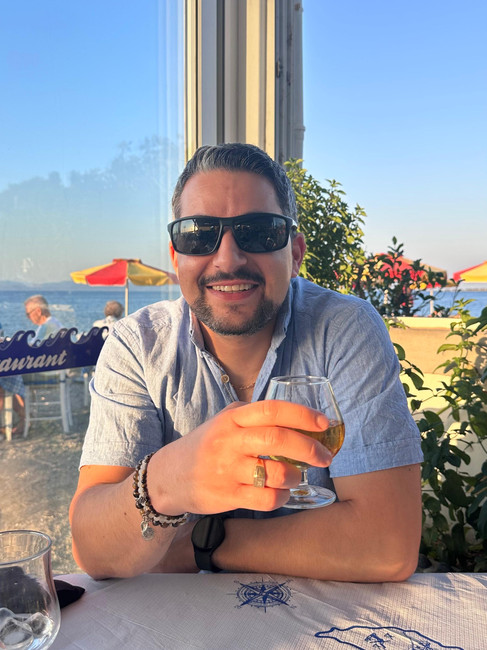 A man wearing sunglasses and a short-sleeved shirt sits at an outdoor table, smiling and holding a drink. The setting is sunny, with a view of the sea, red umbrellas, and greenery in the background.