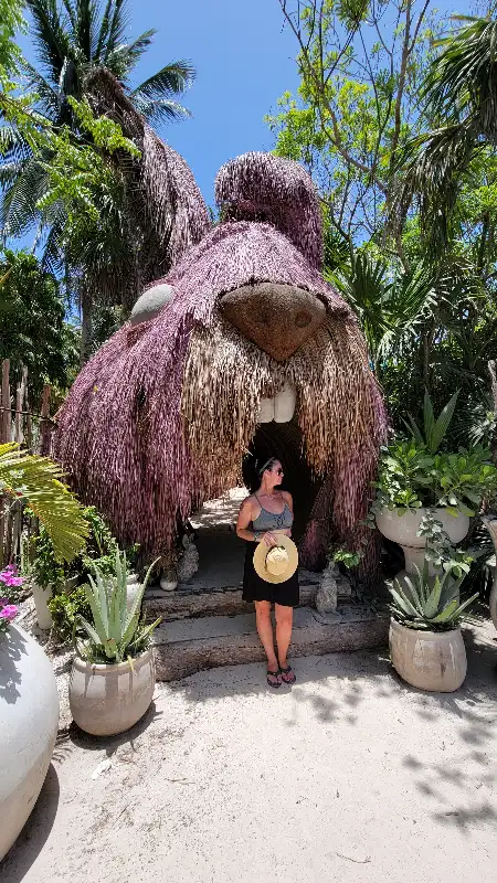 Jenn stands in front of the large, purple, rabbit-shaped entrance sculpture known as Conestesia in Tulum, Mexico, surrounded by tropical plants and potted succulents.