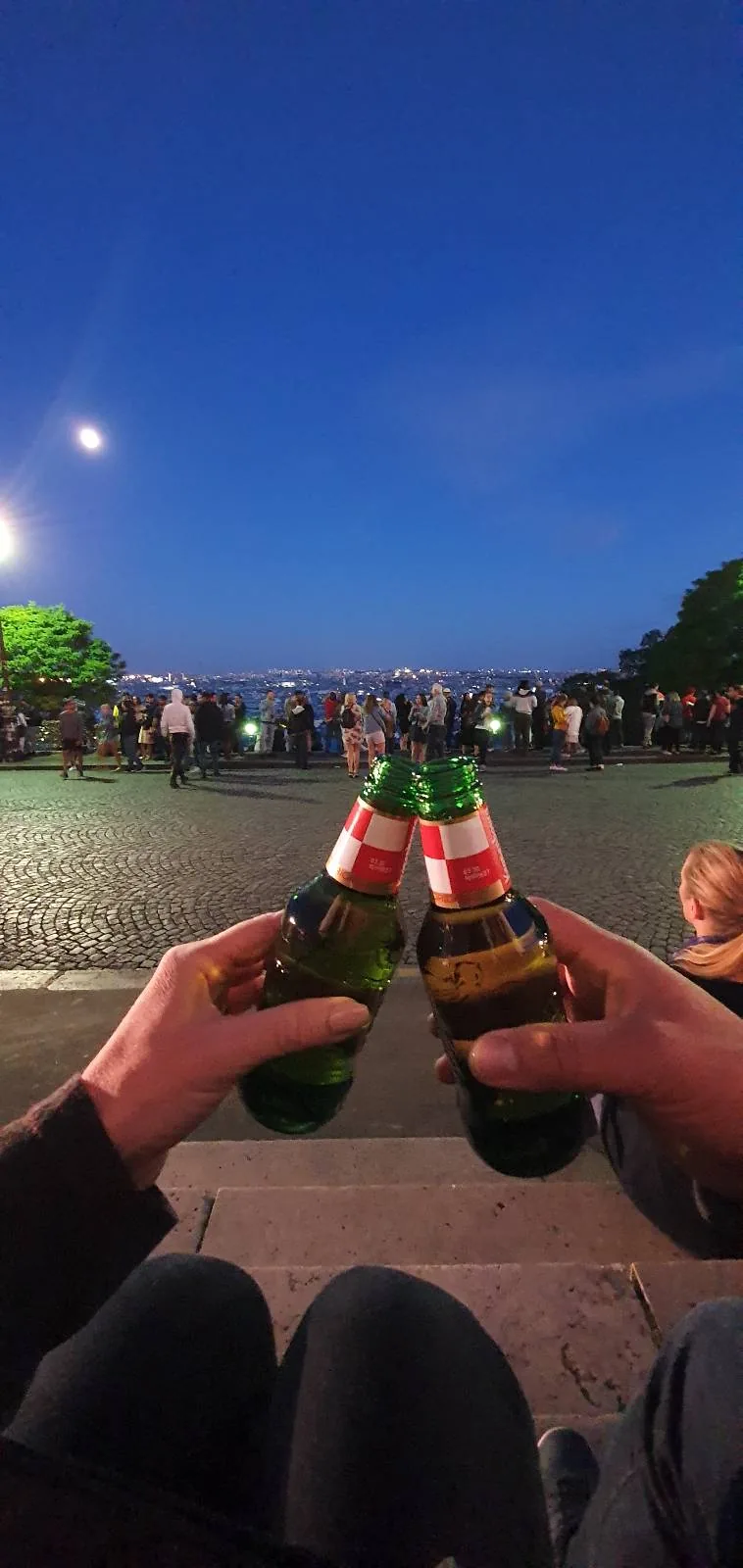 Two people clink beer bottles while sitting on a curb at a nighttime outdoor gathering. A crowd is visible in the background under a clear, moonlit sky.