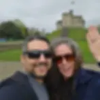 A couple takes a selfie with a historic stone castle in the background, smiling and waving to the camera.