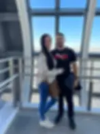 A couple poses smiling in an observation deck. She's in jeans and plaid, he's in a black Puma shirt. Cityscape visible through large windows.