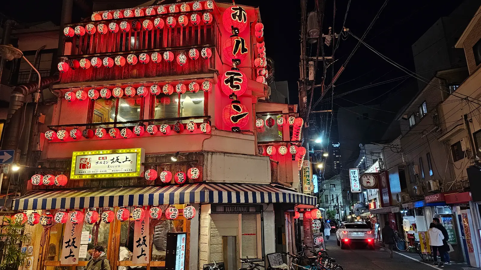 A brightly lit Japanese restaurant at night is decorated with many red lanterns. The street is lively, with people walking and a car parked nearby. Buildings line the street under a dark sky.
