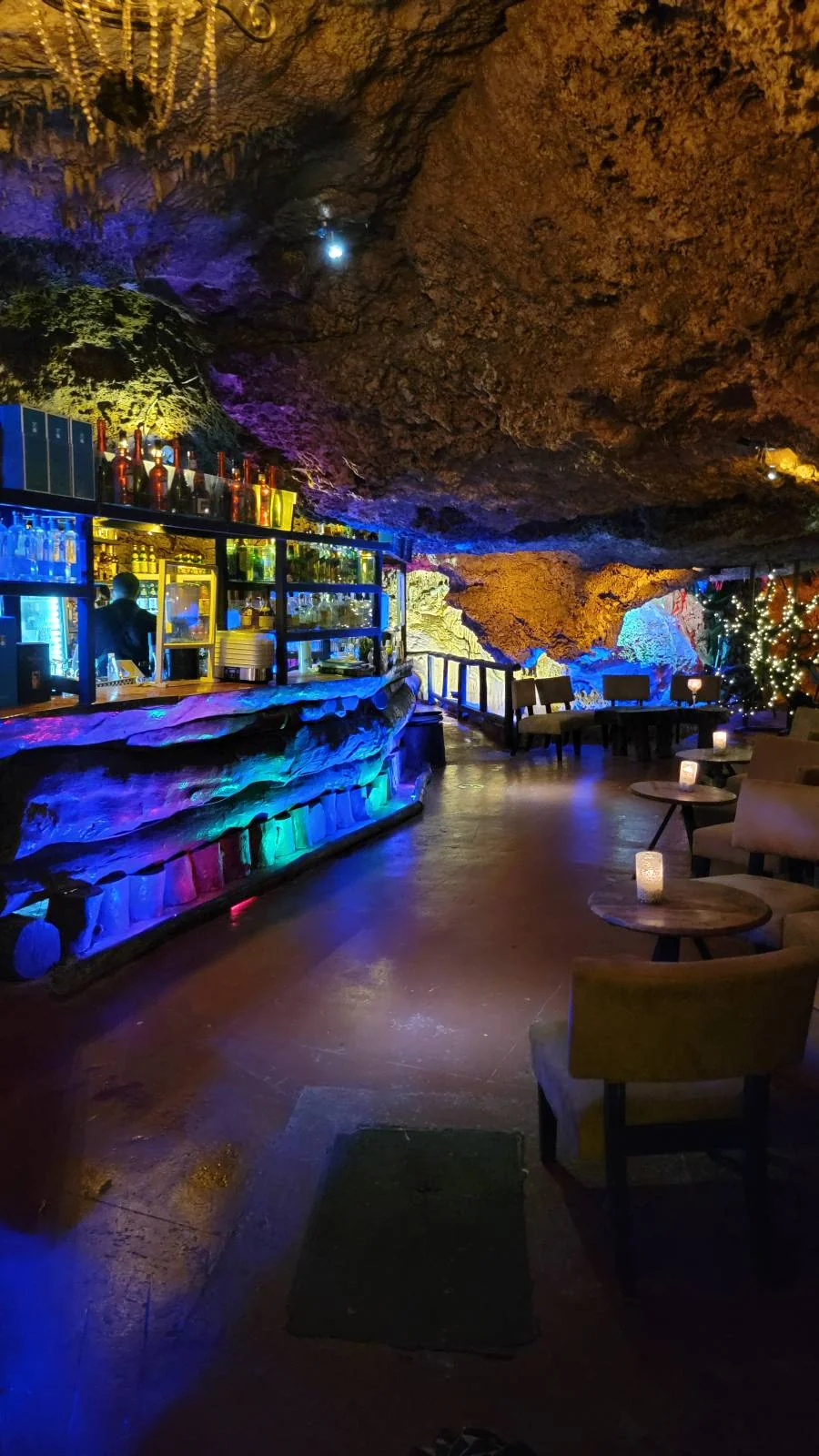 A cave-themed restaurant with colorful lighting. The room features a bar on the left with stools and a counter, and tables with chairs on the right. Illuminated rock formations create a cozy, unique ambiance.