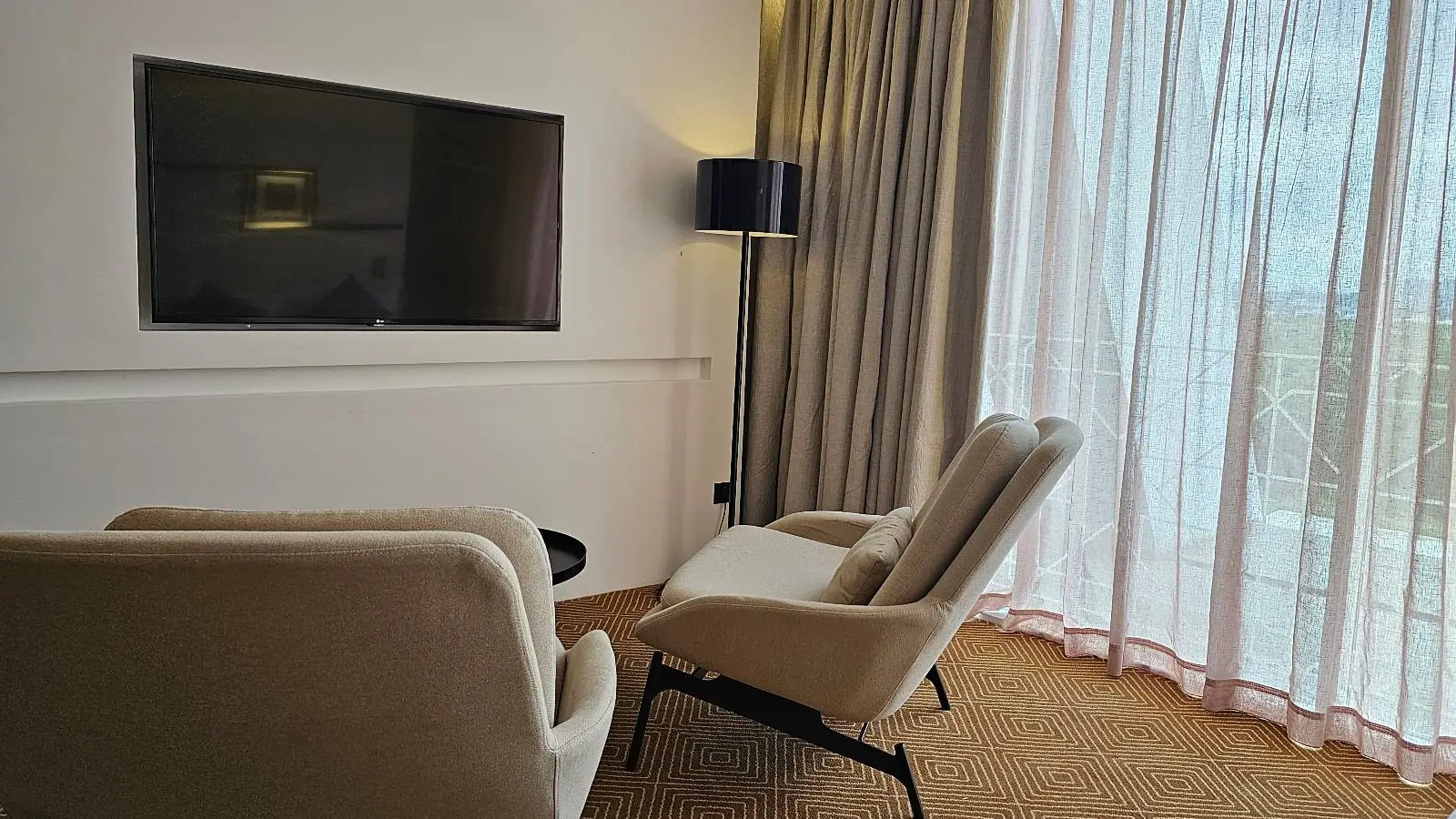 A cozy living room features two beige armchairs facing a large flat-screen TV mounted on the wall. A floor lamp stands near the corner, and sheer curtains allow soft light to filter through, creating a warm and inviting atmosphere.