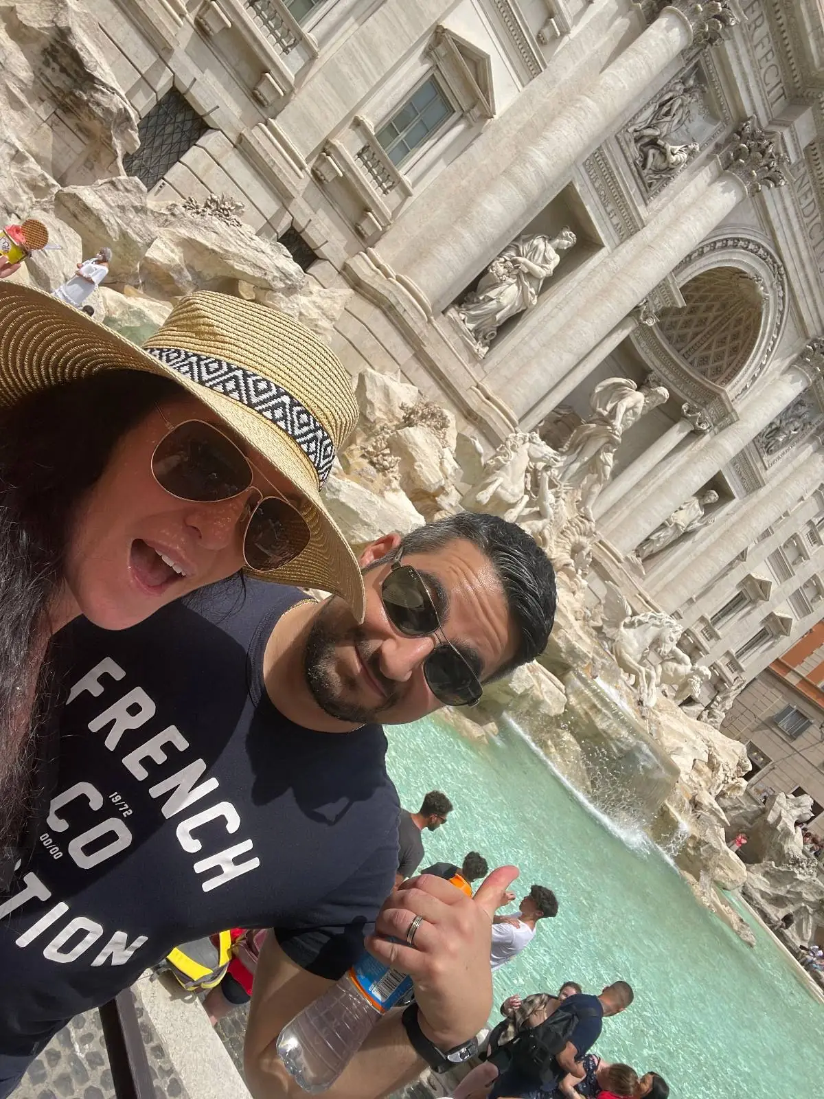 Two people wearing sunglasses pose for a selfie in front of the Trevi Fountain in Rome. The woman wears a sunhat and "French Co Edition" shirt; the man has a beard and black shirt. Other tourists and the fountain's water are visible.