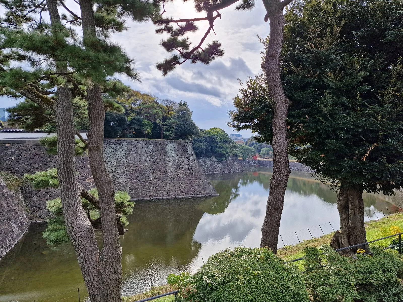 A serene scene of a moat surrounded by stone walls, with trees framing the foreground. The water reflects the cloudy sky and greenery, creating a tranquil atmosphere.