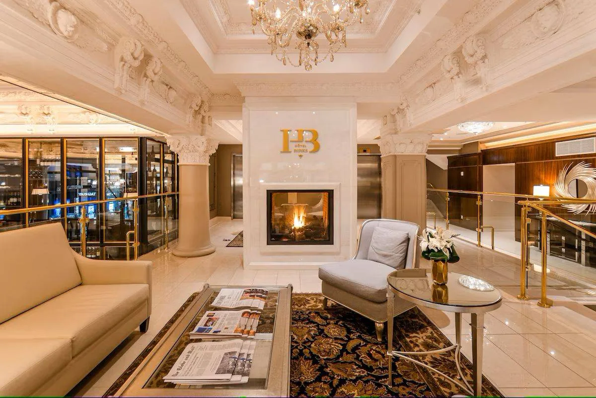 Elegant hotel lobby with chandelier, fireplace, and beige seating. Newspapers on table, ornate ceiling details, and "HB Hotel" sign above fire.