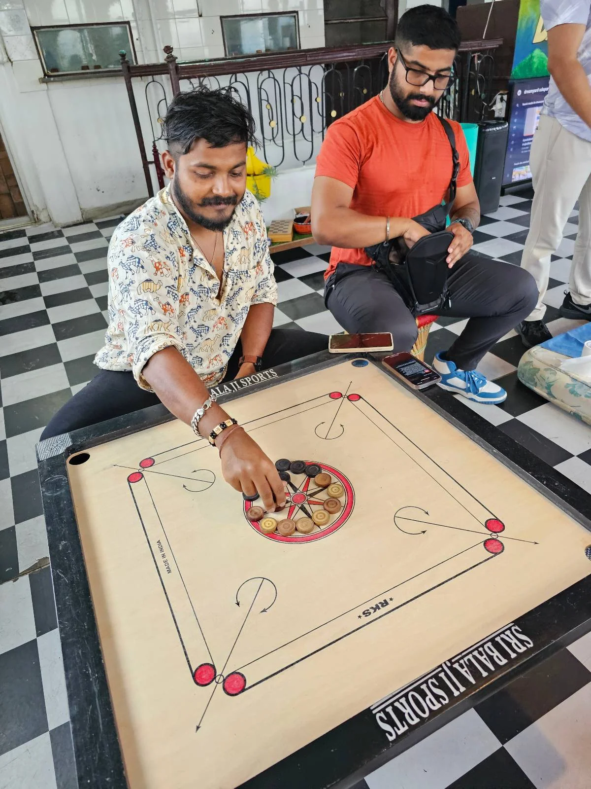Two men sit on the floor playing carrom on a carrom board. One man in a patterned shirt is about to flick a striker, while the other in a red shirt watches and holds a bag. The floor has a black-and-white checkered pattern.