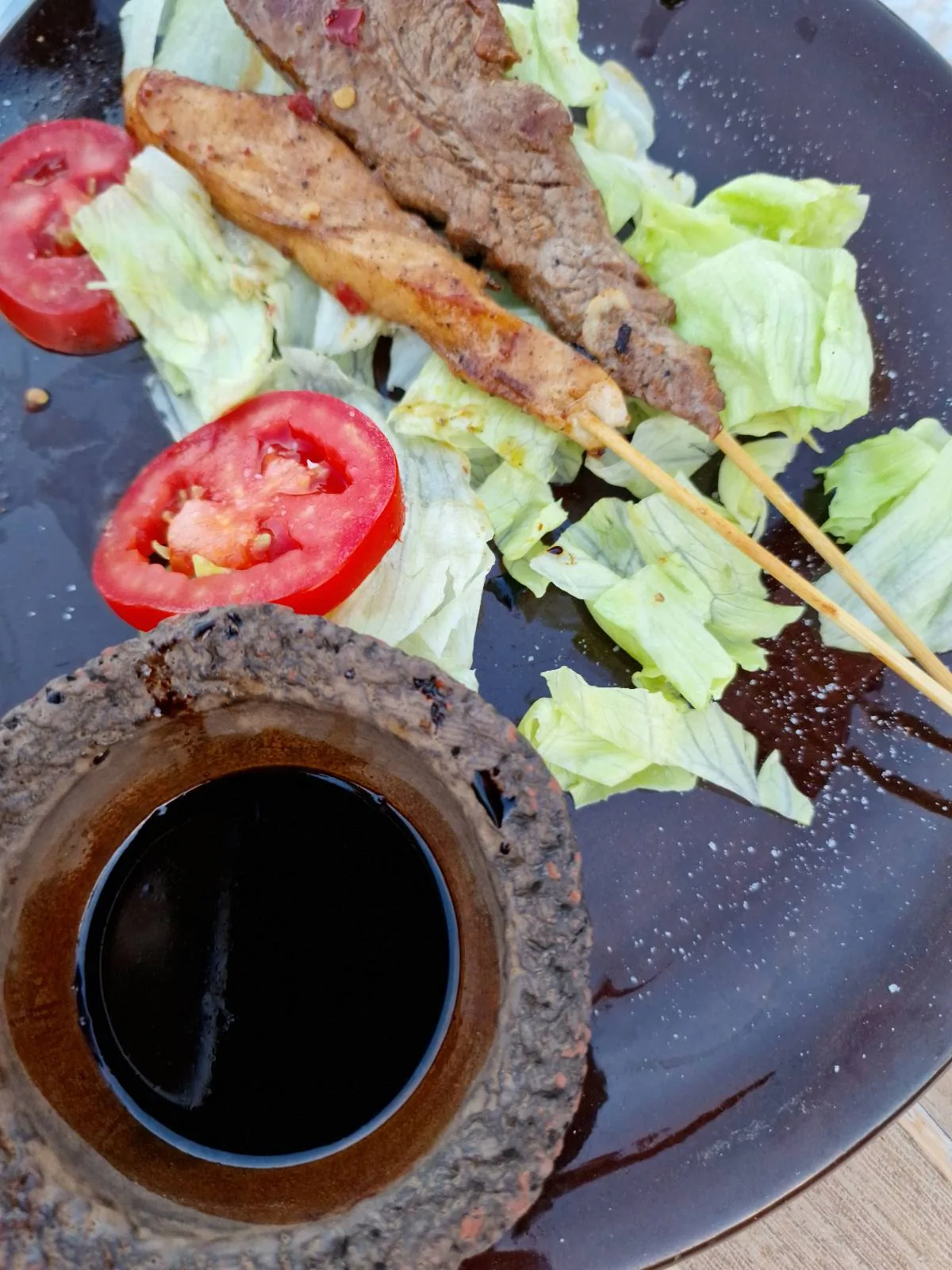 Skewered meat with sliced tomatoes and lettuce on a plate, accompanied by a small bowl of dark dipping sauce.