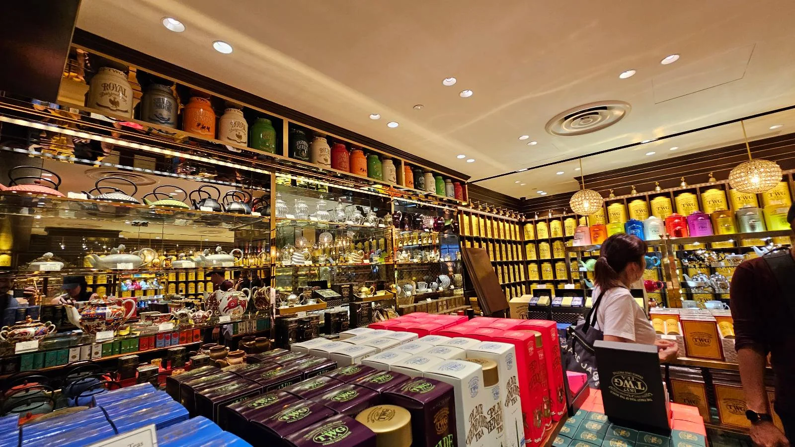 A vibrant tea shop with shelves full of tea canisters, teapots, and accessories. Various boxes are stacked on tables, and colorful tea tins line the walls. Customers browse the offerings in the warmly lit store.