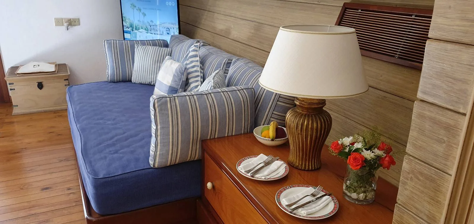 A cozy room features a blue sofa with striped pillows against a wooden wall. A wooden table beside it holds a lamp, a fruit bowl, and a vase with red and white flowers. The floor is wooden, and a small box sits in the corner.