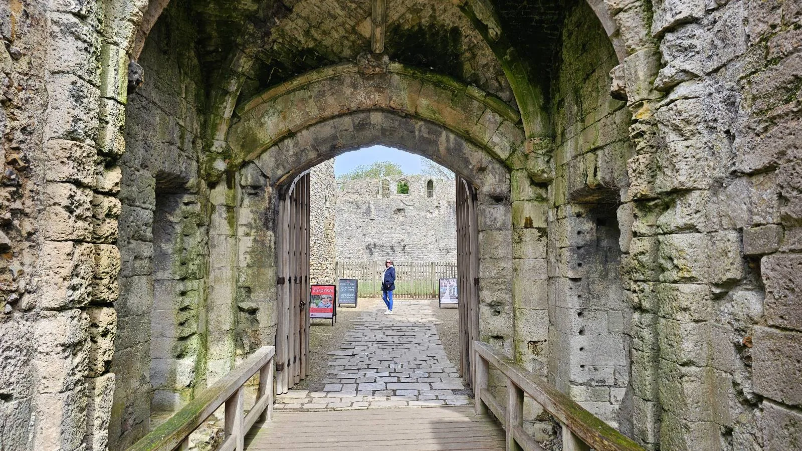 Stone archway entrance of an old castle or fortress with mossy walls, wooden doors, and cobblestone ground. A person stands outside in the courtyard, with a blue sky and stone buildings visible in the background.