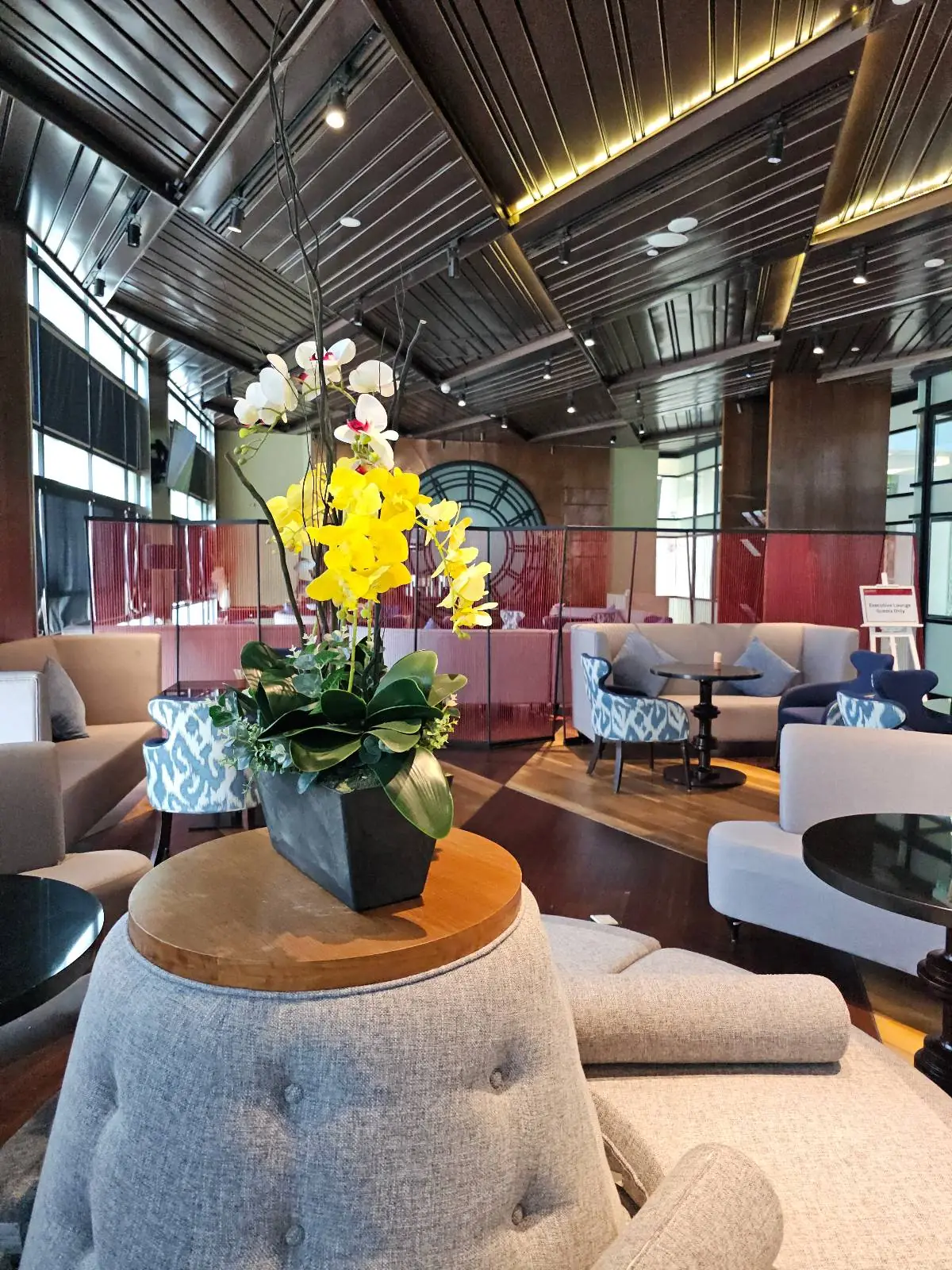 A modern lounge features elegant seating with plush gray chairs and a central table adorned with a vibrant bouquet of yellow and white flowers. The room is spacious, with large windows and a high ceiling, creating an inviting atmosphere.