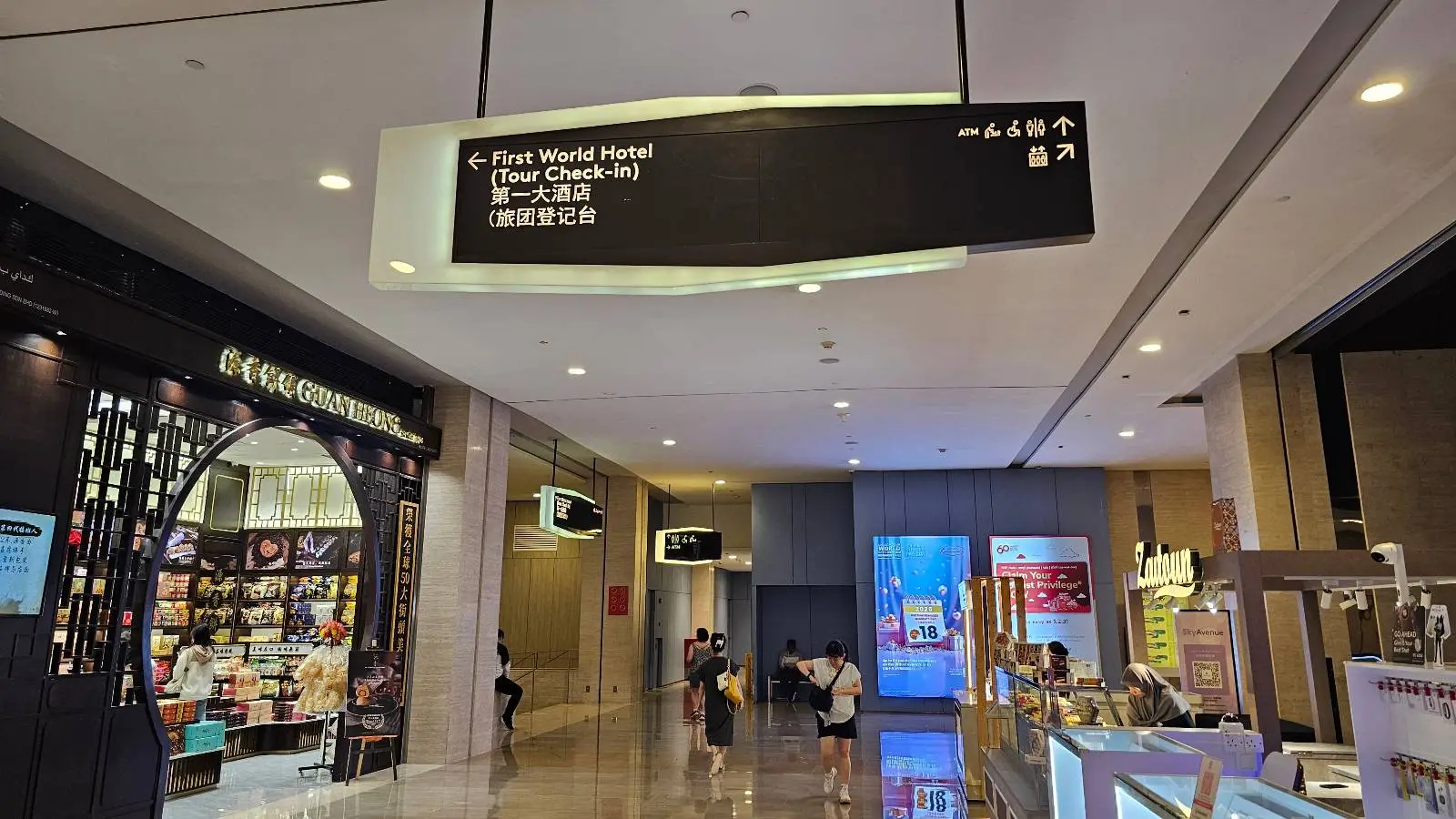 A shopping mall corridor with stores on both sides, a few people sitting, and a black overhead sign in English and Chinese displaying directions to various locations.