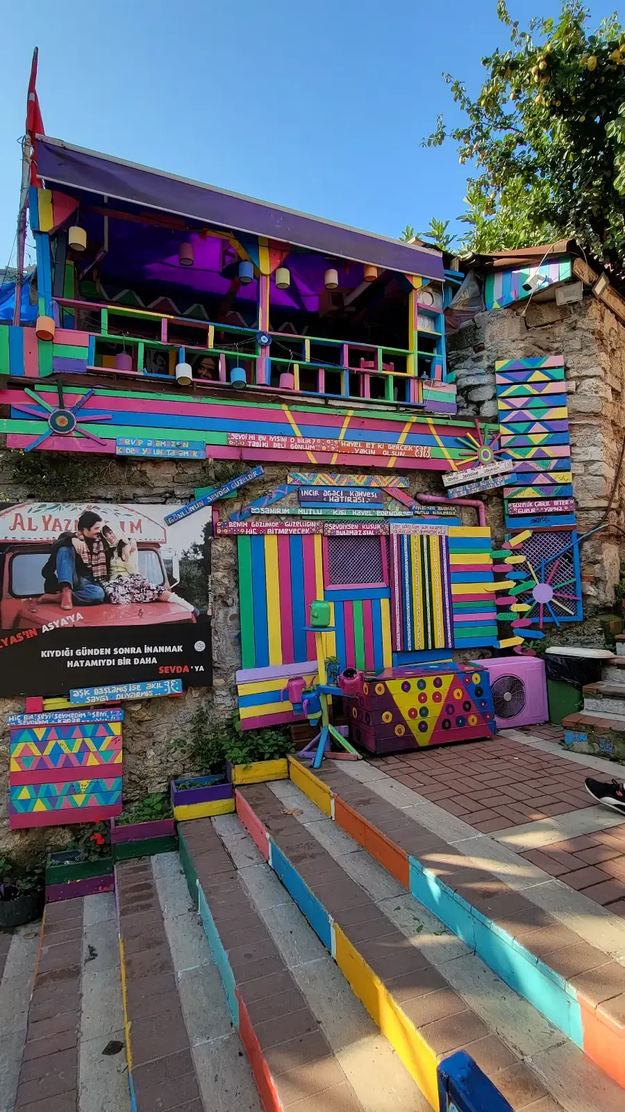 A colorful building with vibrant rainbow-painted walls, doors, and steps, featuring a balcony with purple awning. Murals and geometric patterns cover the exterior, creating a lively, artistic atmosphere under a clear blue sky.