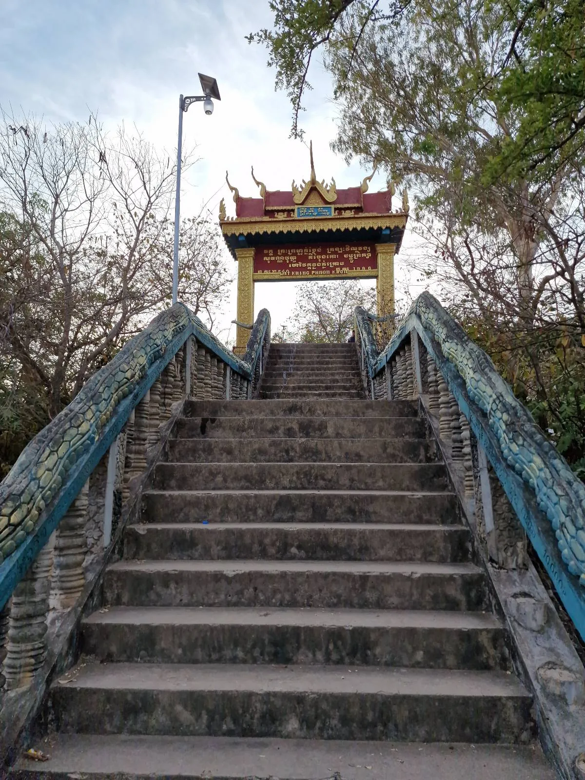 Stone stairs with dragon railings lead up to a colorful, ornate gateway at the top, surrounded by leafy trees and a cloudy sky in the background.