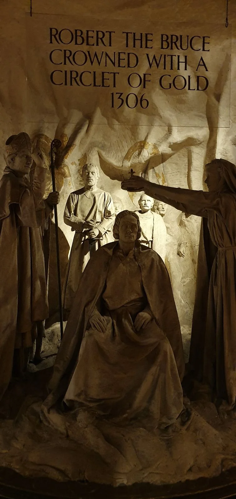 Sculptural scene of Robert the Bruce being crowned in 1306, with figures presenting a gold circlet inside a historic chamber.