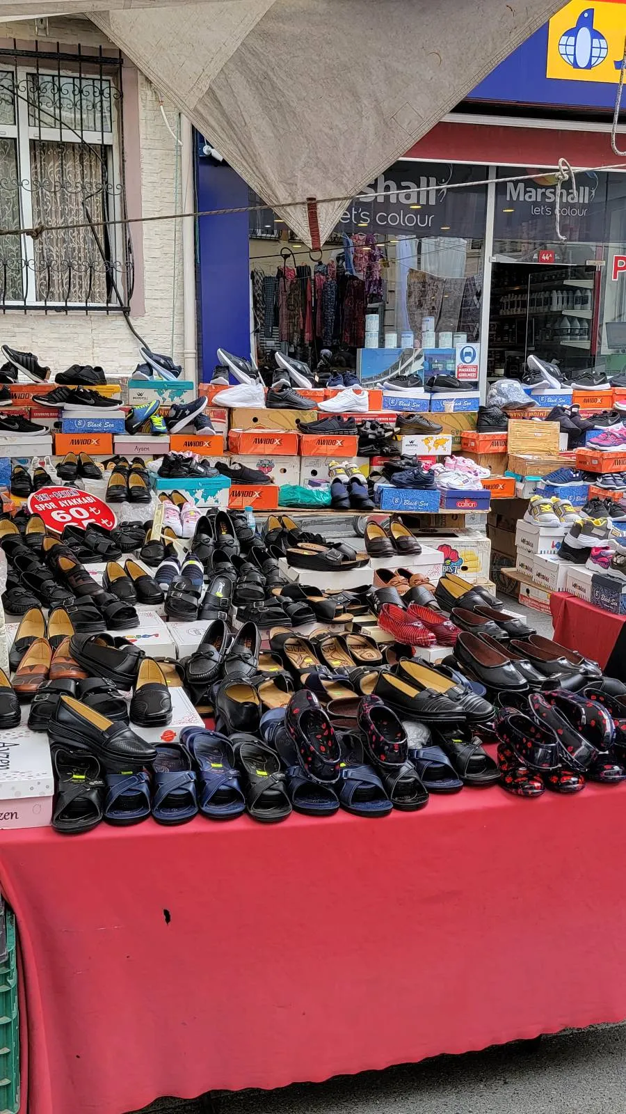 A market stall displays various shoes, including sandals, dress shoes, and sneakers, on a red tablecloth under an umbrella.