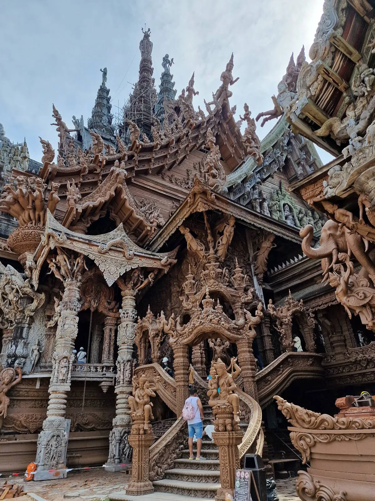 A person ascending a staircase leading to an intricately carved wooden temple with elaborate sculptures and pointed spires. The architecture is detailed and ornate, set against a cloudy sky.