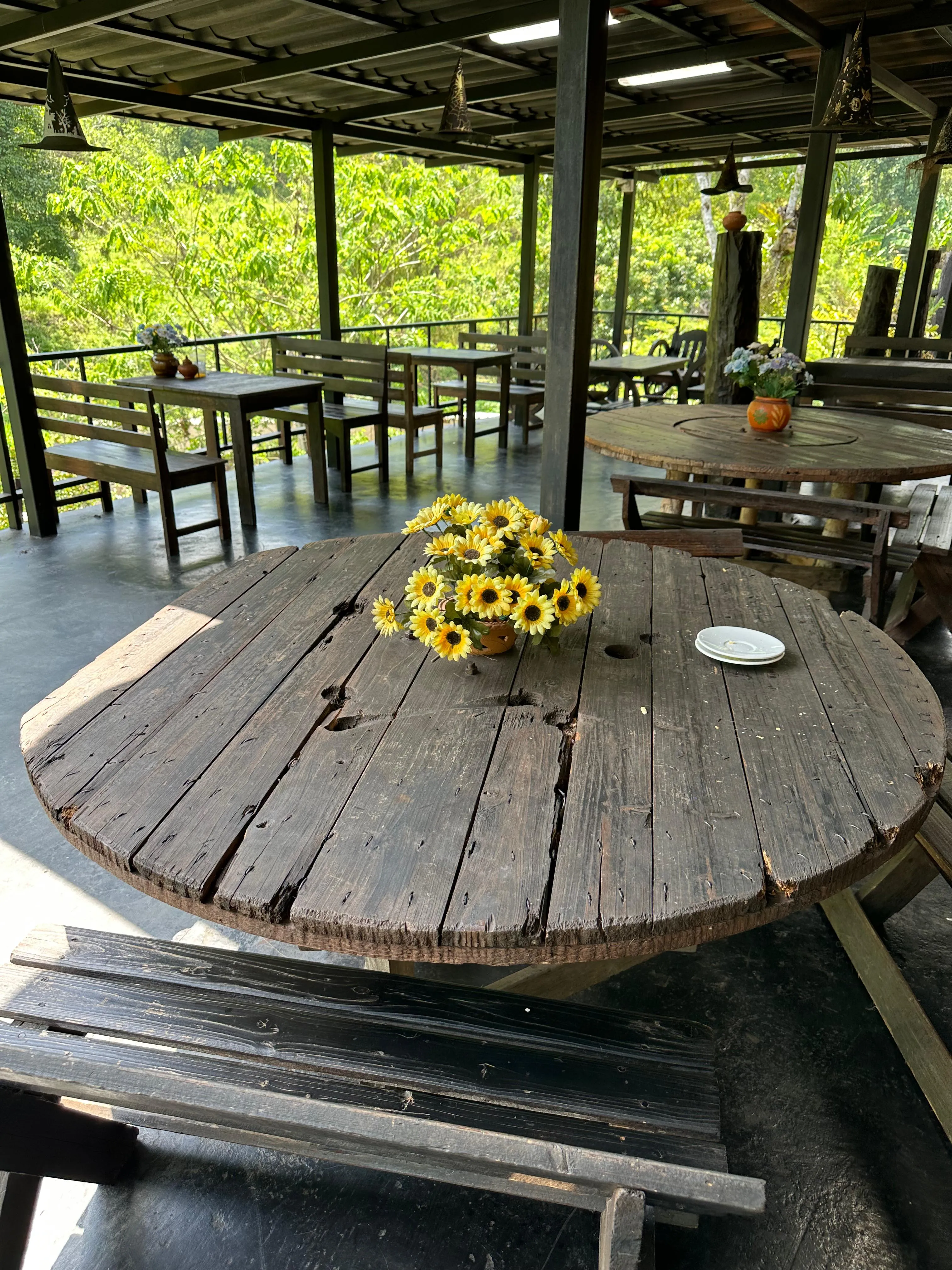A rustic wooden round table with a bouquet of yellow flowers in a vase at its center, surrounded by benches in an open-air, covered seating area with greenery visible outside.