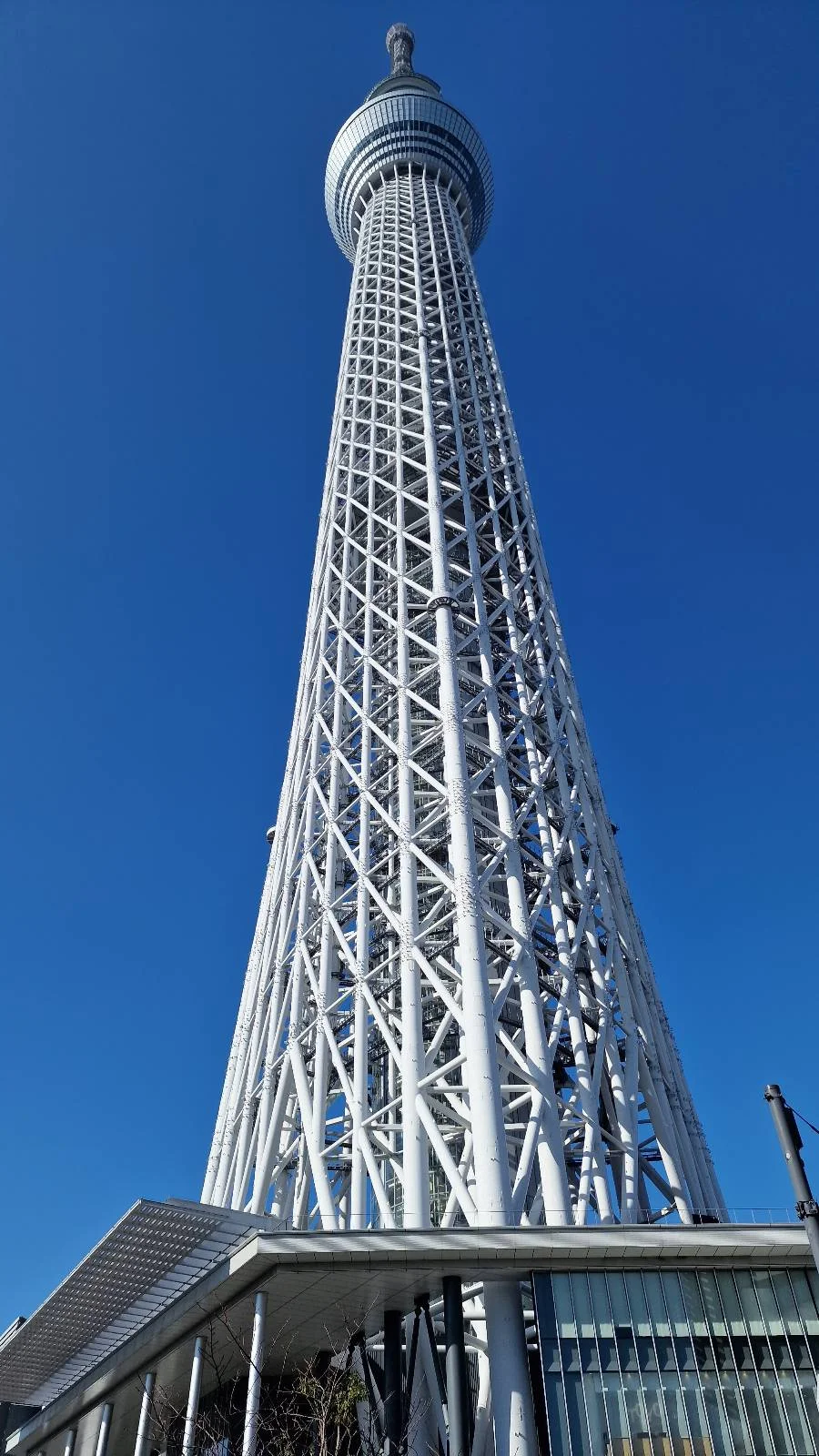 A towering, white lattice structure known as the Tokyo Skytree stretches skyward against a clear blue sky. Seen from below, the angle accentuates its height and sleek design. The observation deck and antenna are visible at the top.