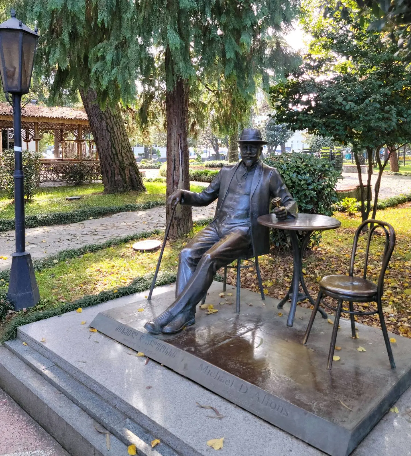Bronze statue of sitting man on a raised plinth, with arm resting on a table accompanied by an empty bronze chair to its right.