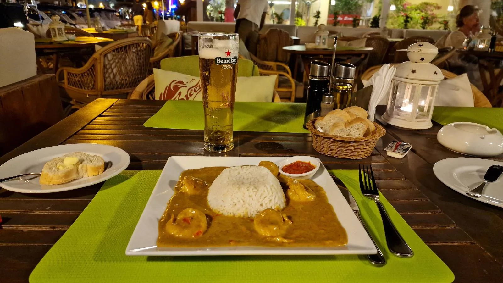 A square white plate with rice and curry sits on a wooden table set with a glass of beer, a basket of bread, and a candle lantern at an outdoor dining area.