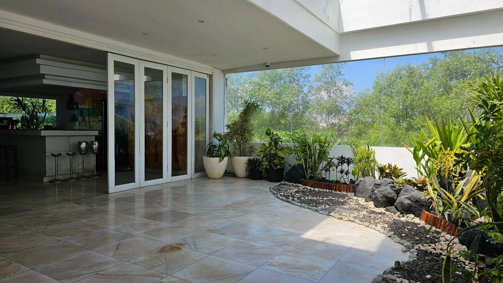 Spacious indoor area with large sliding glass doors opening to a lush garden view. The room features a tiled floor and includes various potted plants and decorative stones near the entrance, enhancing the natural ambiance.