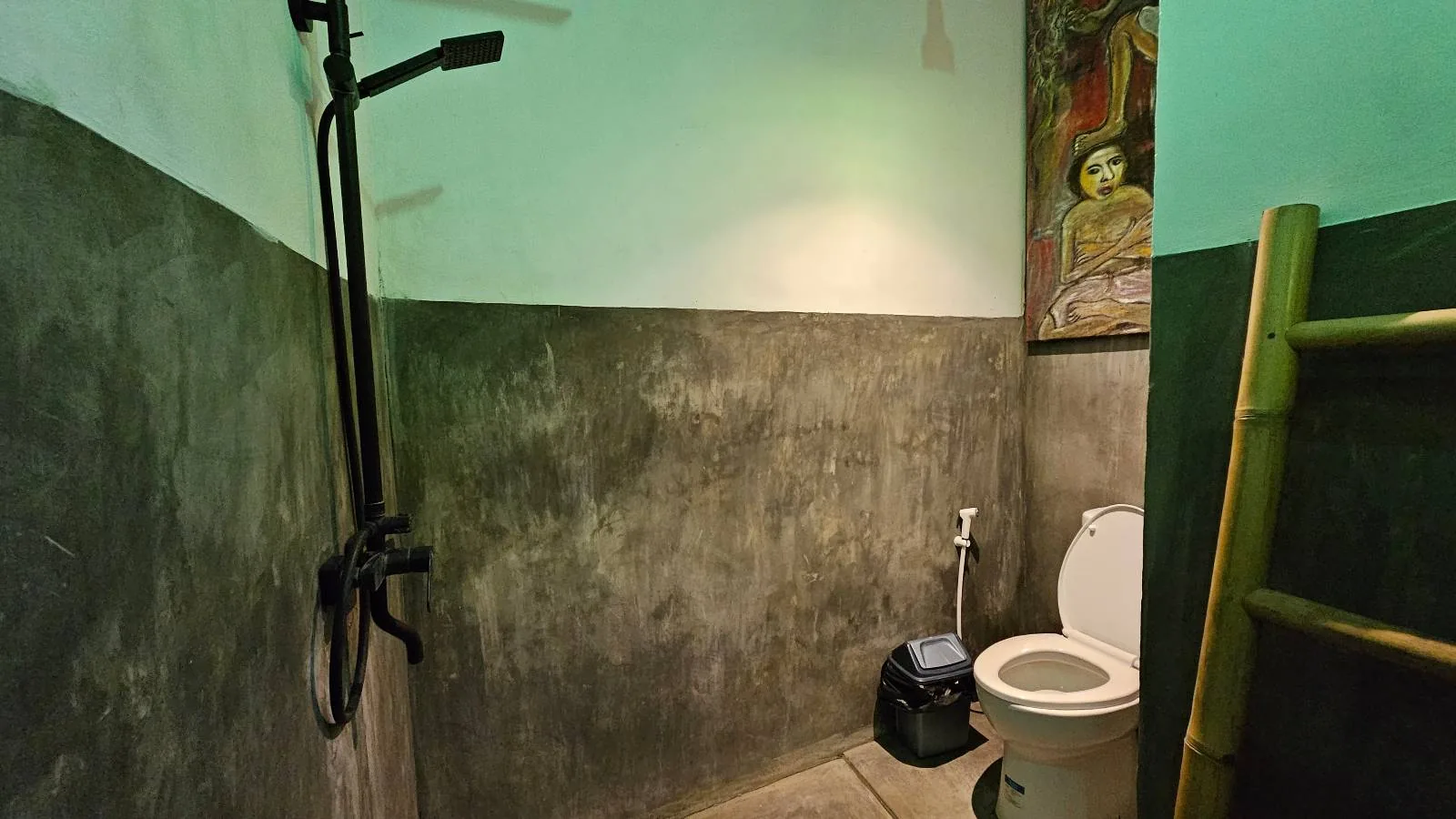 A bathroom with greenish walls features a toilet and an overhead shower with a black fixture. A bamboo ladder leans on one wall, and an artwork is partially visible above the toilet. The lighting gives the room a muted, ambient glow.