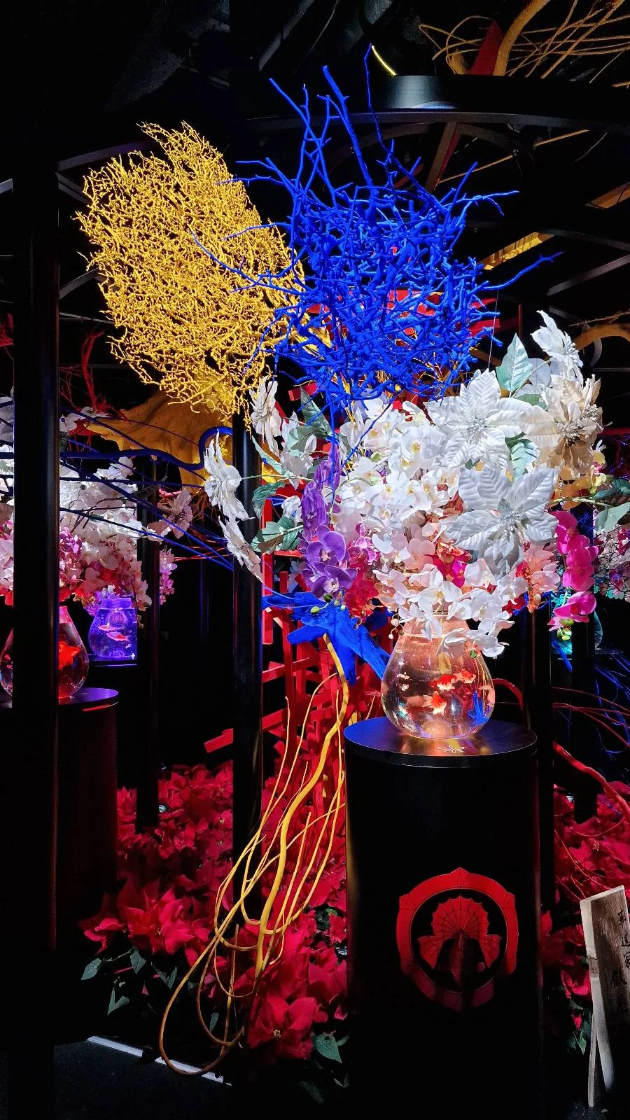 A vibrant display of colorful, abstract floral arrangements in a dimly lit room. The composition features bright yellows, blues, and purples with varied textures, placed in glass vases on pedestals, creating an artistic and dramatic visual effect.