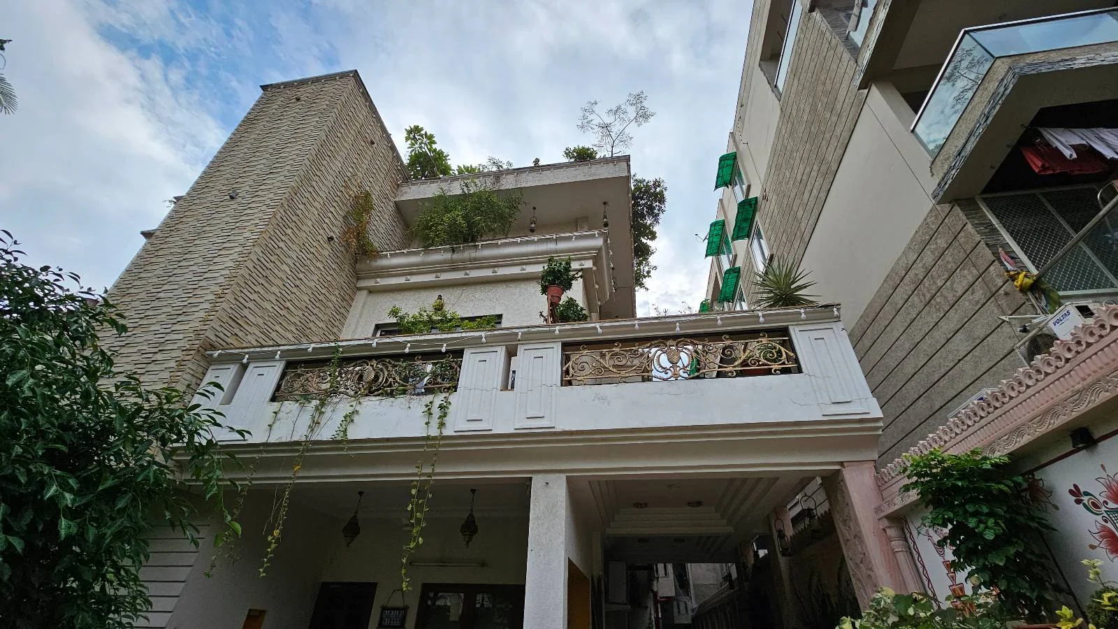 A low-angle view of a multi-story residential building with a white balcony, green plants, and neighboring buildings on either side, set against a partly cloudy sky.