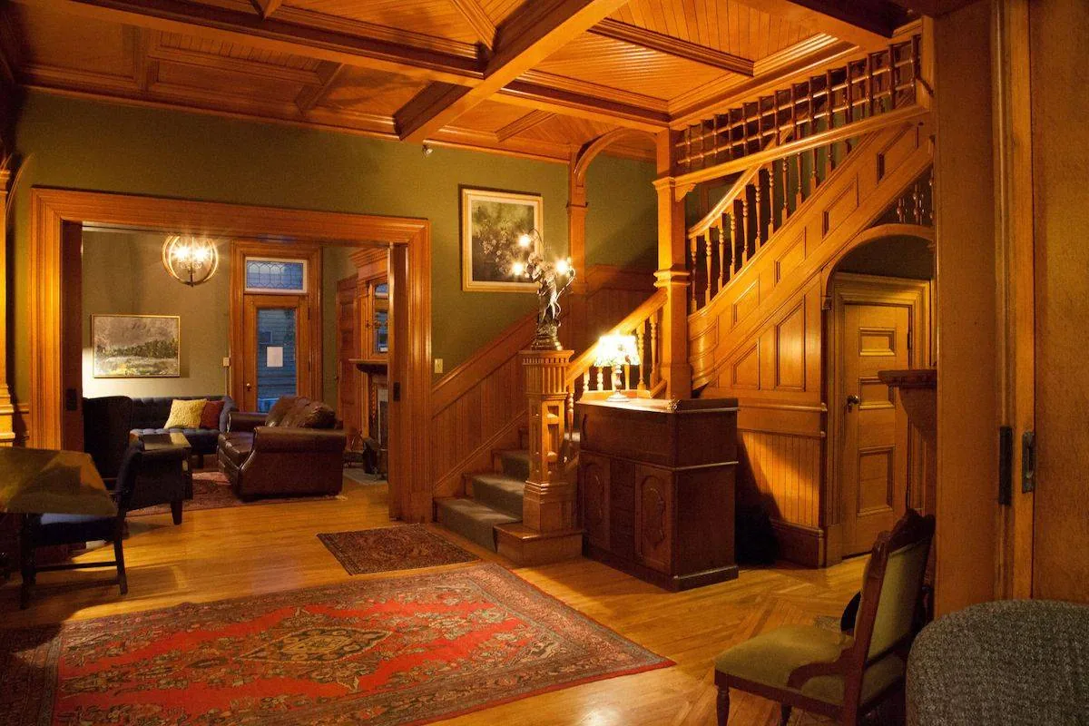 A warmly lit interior with wooden walls and ceiling, featuring a grand staircase. There's an area rug on the wooden floor, a chandelier, and several pieces of furniture, including chairs and a cabinet. A painting hangs on the green wall.
