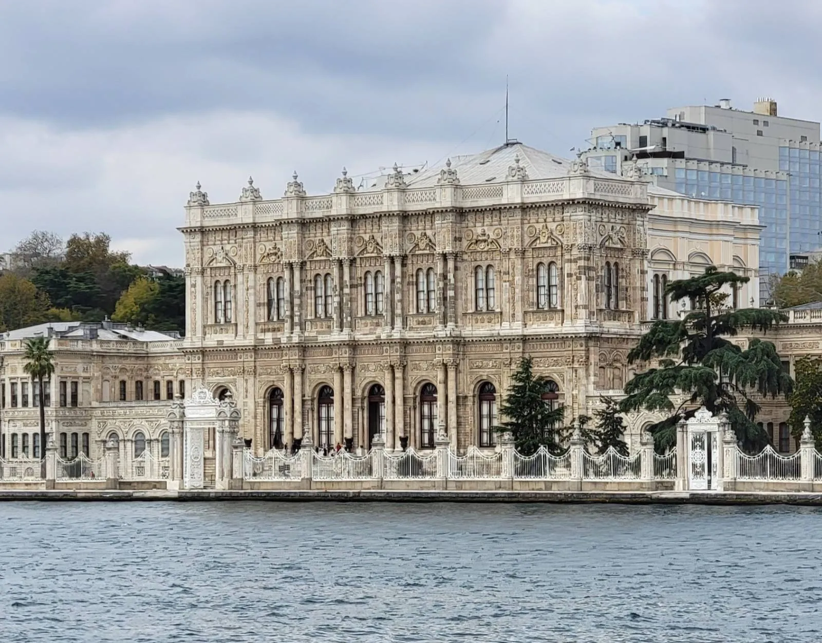 A grand, ornate building with classical architecture stands by the waterfront under a cloudy sky, featuring large windows, columns, and intricate details. Trees and modern buildings are visible in the background.