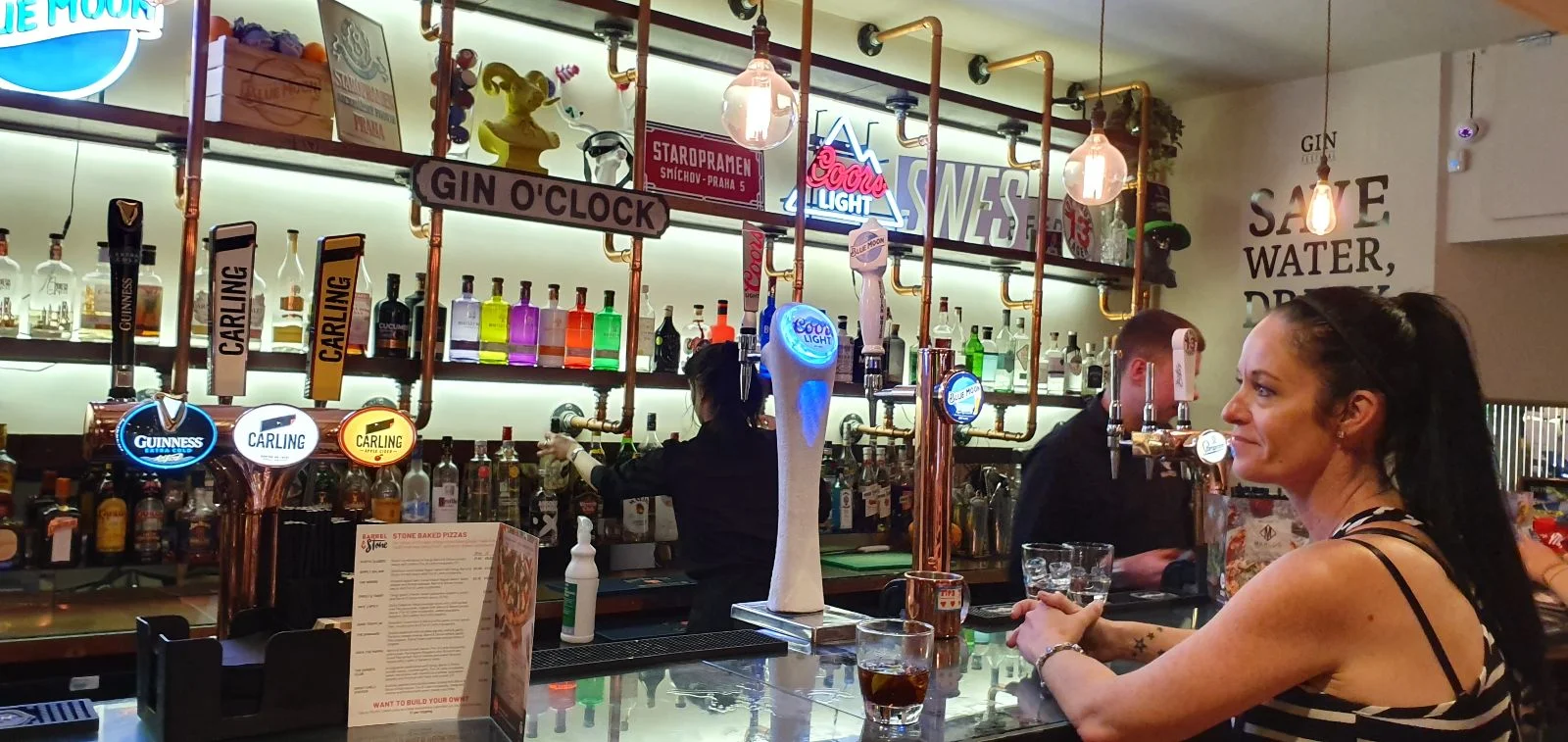 Woman at a lively bar counter with colorful bottles, neon signs, and taps. Bartenders serve drinks; text reads "GIN O'CLOCK" and "SAVE WATER."