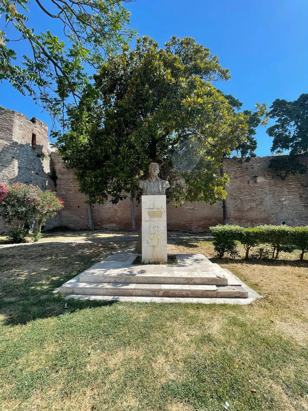 A bust on a pedestal stands in a grassy area, surrounded by trimmed bushes and trees, with a stone wall and bright blue sky in the background.