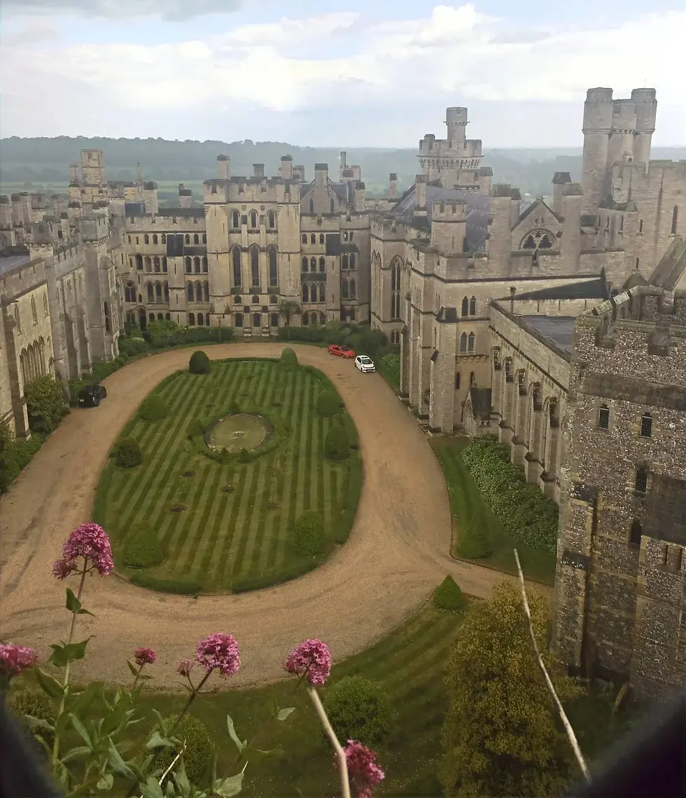 Castle courtyard with manicured lawns, a circular driveway, and parked cars. Flowers in foreground, cloudy sky above the historic stone building.