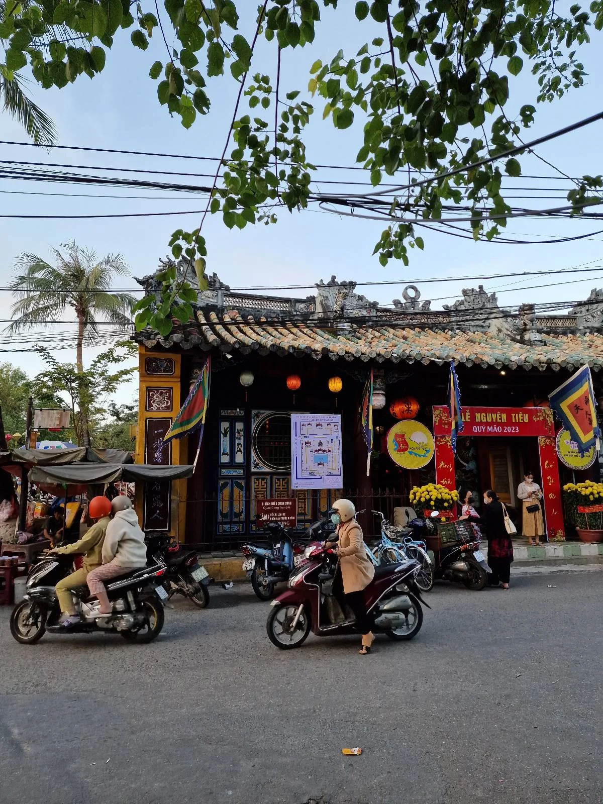 Street scene with people on motorbikes in front of a traditional building, decorated with colorful banners and lanterns. Palm trees and overhead wires are visible in the background.