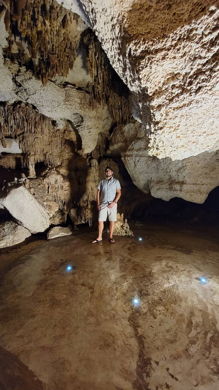 A person stands in a cave, surrounded by rock formations. They are wearing a light-colored shirt and shorts, with sunglasses. Small lights are embedded in the ground around them, illuminating the cave interior.