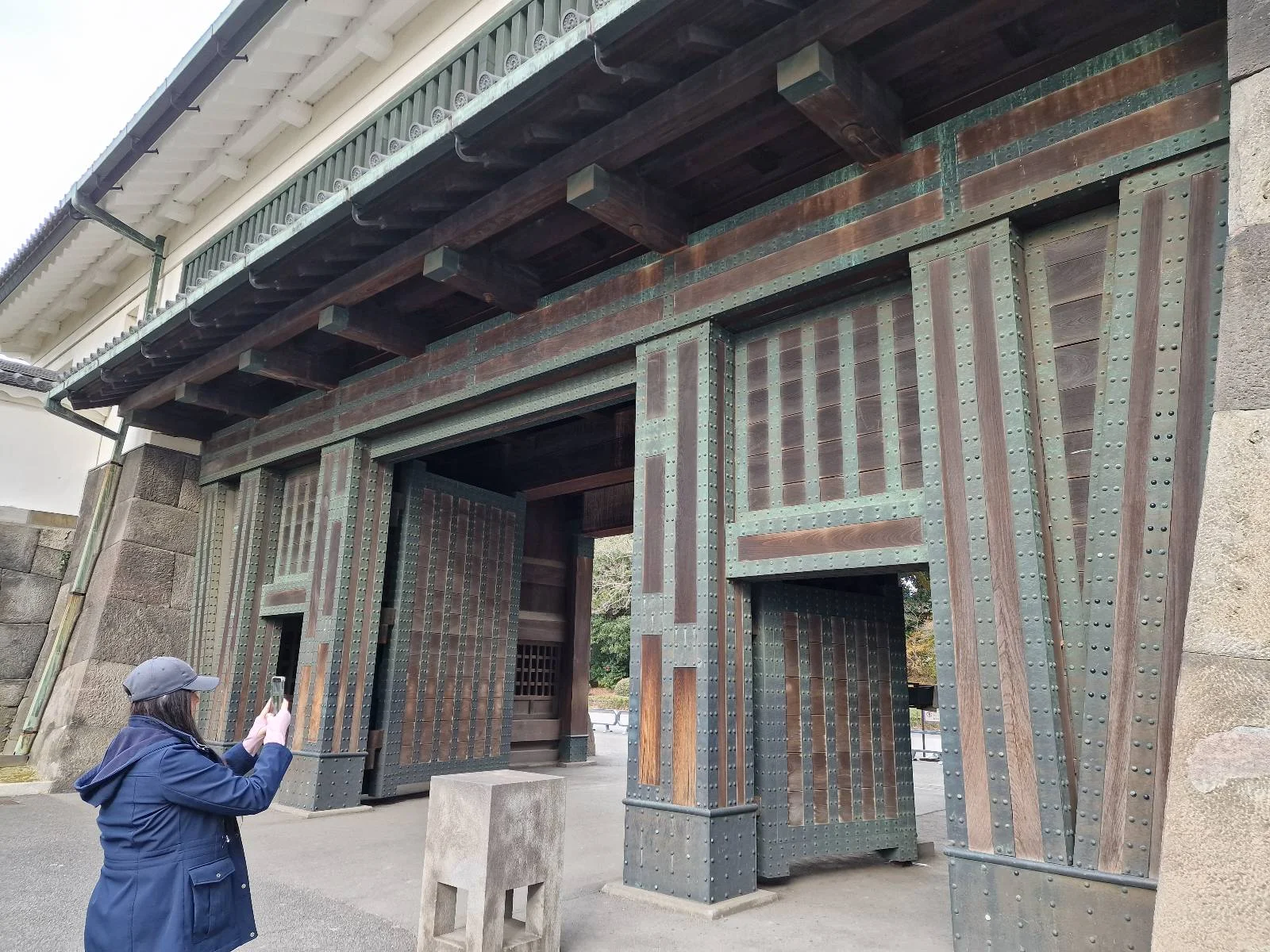 A person in a blue jacket and cap takes a photo of a large, ornate wooden gate with intricate patterns. The structure features a combination of wood and metal elements. Stone pillars are visible in the foreground.