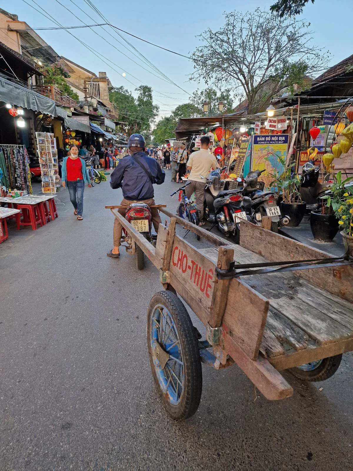 A bustling street market scene with people walking and browsing colorful goods. A wooden cart with a bicycle attached is in the foreground. Stalls with vibrant textiles, plants, and various items line both sides of the street.