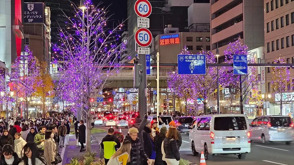 Crowded city street at night with purple-lit trees, traffic, and many pedestrians. Japanese signs visible, festive and lively atmosphere.