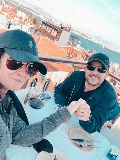 Two people wearing sunglasses and caps hold hands at an outdoor cafe table, with a drink and plates, overlooking a cityscape and body of water in the background.