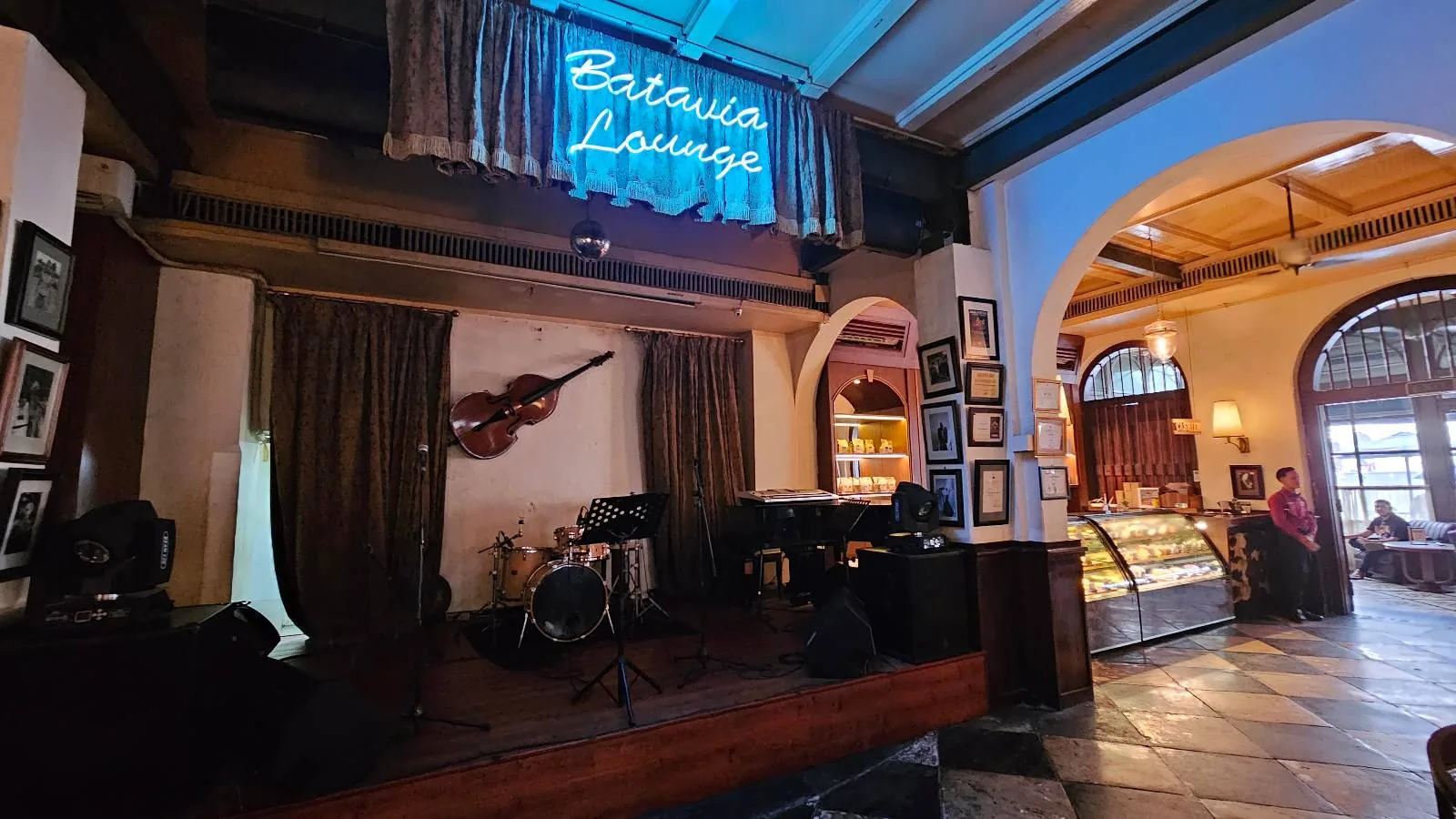 A cozy lounge with a small stage featuring musical instruments and a microphone setup. A neon sign reads "Batavia Lounge" above. There are framed pictures on the walls and a pastry display in the background.