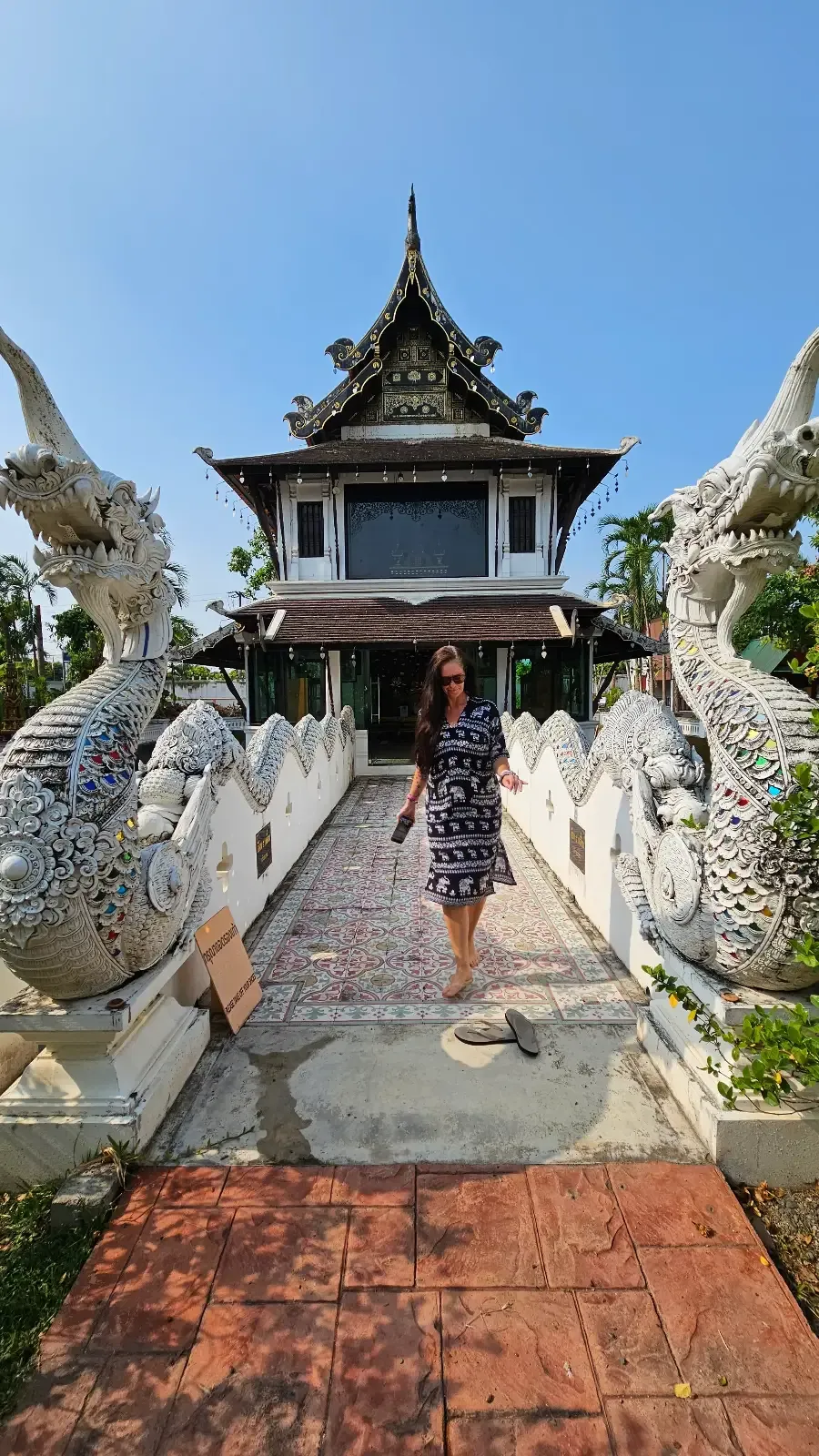 A person in a striped dress walks across a decorative bridge flanked by white dragon statues toward a traditional, ornate building with a tiered roof under a blue sky.