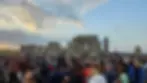 A large crowd gathers at Stonehenge during sunset. People socialize and celebrate, some holding colorful flags. The sky is partly cloudy.