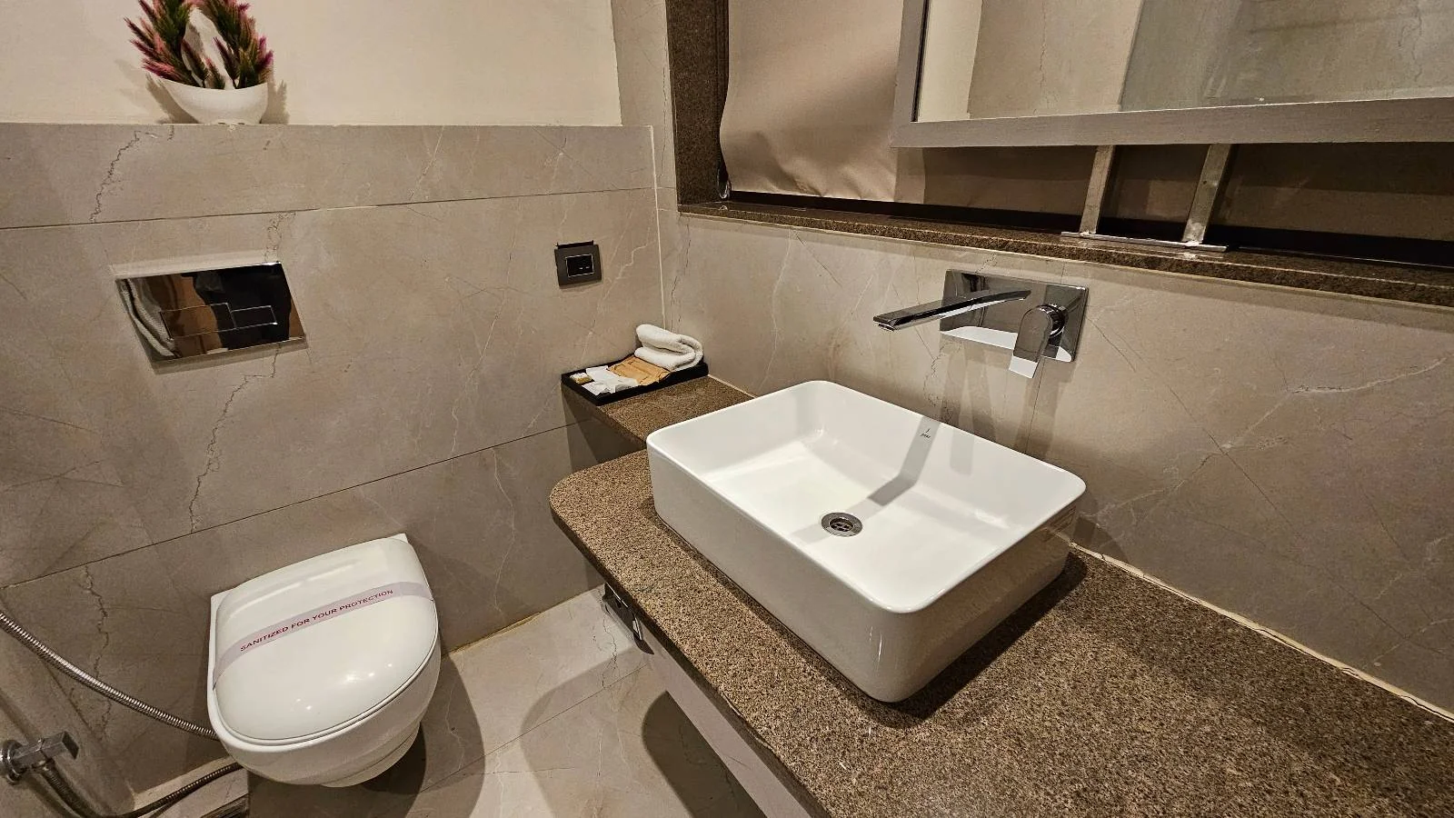 Modern bathroom with a wall-mounted toilet, rectangular sink on a brown countertop, wall-mounted faucet, mirror cabinet, and a small plant on a shelf above the toilet. Towels are neatly placed by the sink.
