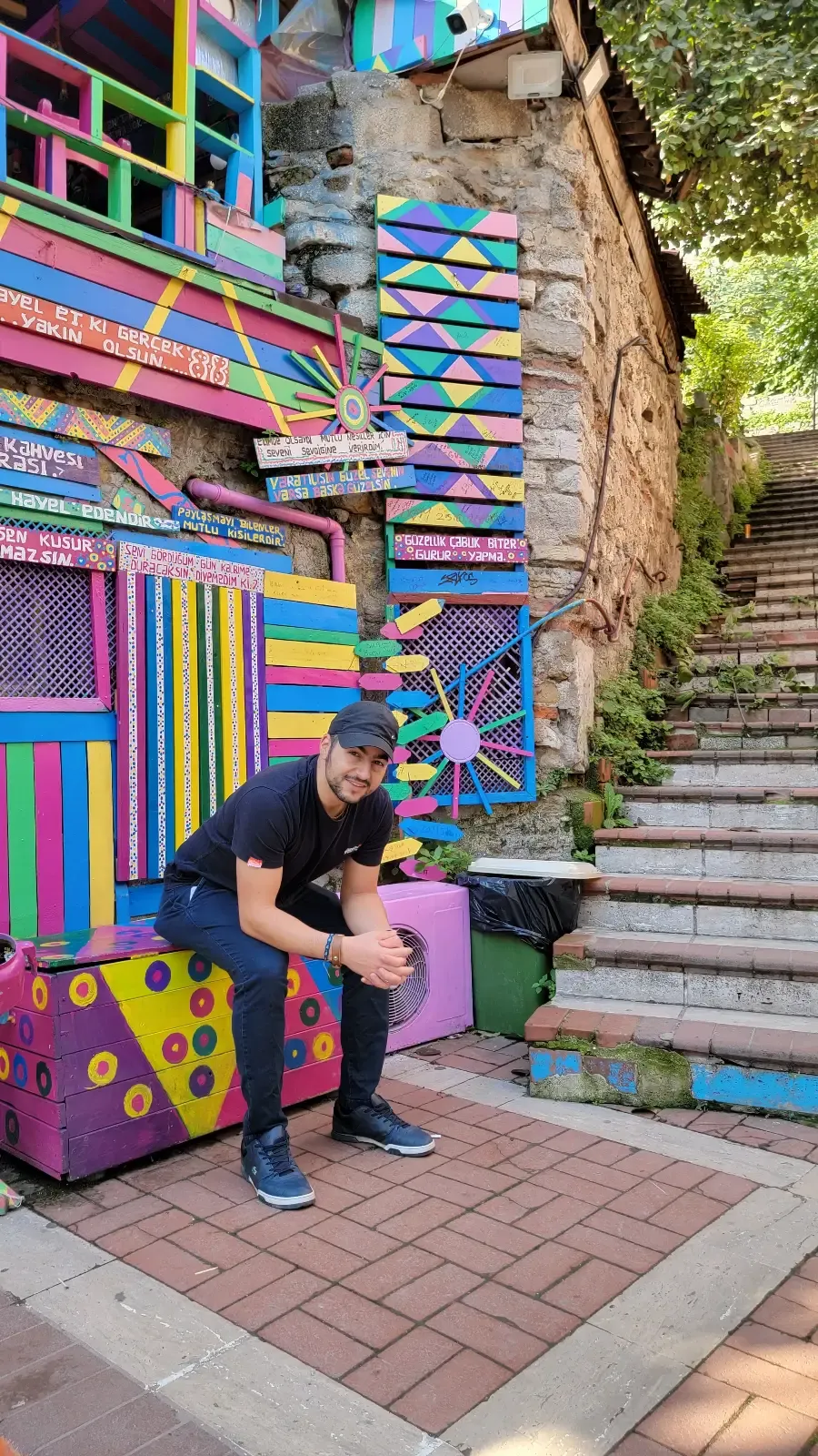 A man sits on a colorful, painted bench in front of a vibrantly decorated building, with patterned walls and doors. Stone steps and greenery are visible to the right.