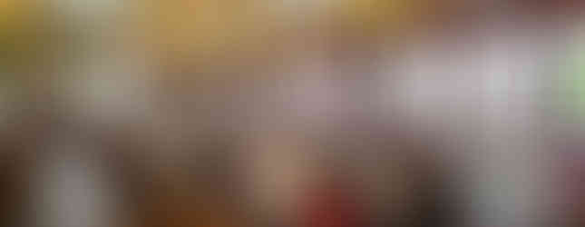 A large group of people sits around a circular table in a spacious restaurant with high ceilings and many windows. The room is well-lit with hanging lamps, and other tables are visible in the background. Some people are engaged in conversation.