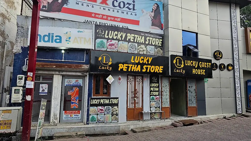 Storefront of Lucky Petha Store and Guest House. Signs feature sweets and a bright yellow-black color scheme. An ATM is nearby.
