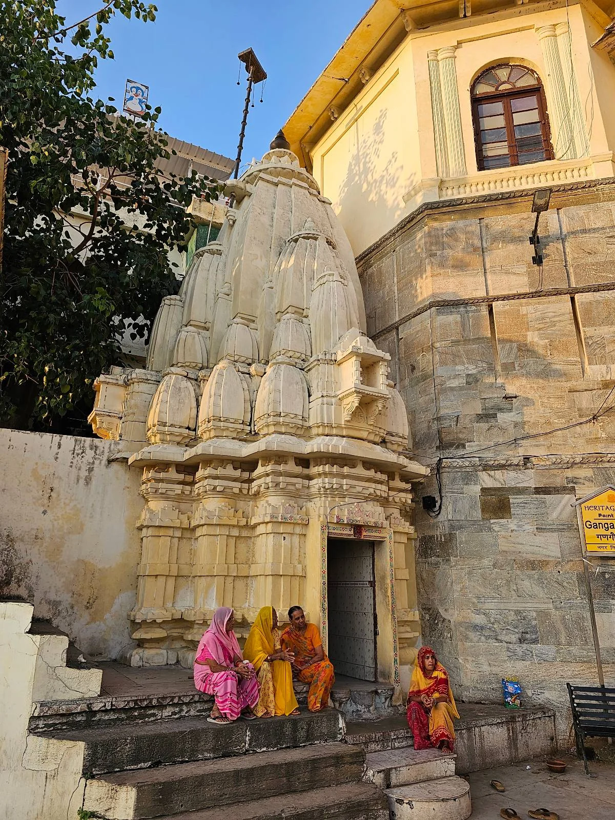 Four people in colorful traditional clothes sit on steps outside an ornate, cream-colored temple with intricate carvings, next to a stone wall and under a clear blue sky.